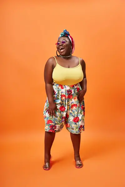 Веселый африканский американец плюс размер женщина в цветочной юбке и солнцезащитных очках позирует на оранжевом фоне — стоковое фото