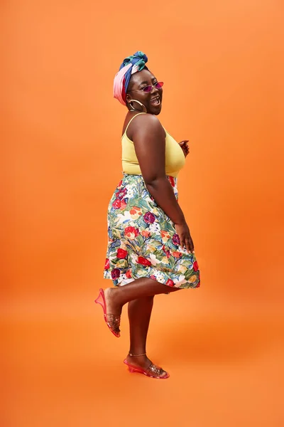 Élégant afro-américain plus taille femme avec jupe florale et lunettes de soleil debout sur orange — Photo de stock