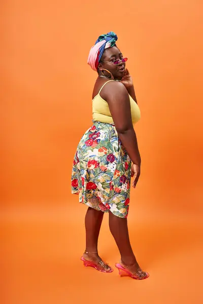 Elegante afro-americana plus size mulher com saia floral e óculos de sol posando em pano de fundo laranja — Fotografia de Stock
