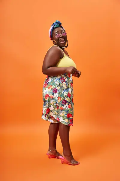 Изумленный африканский американец плюс размер женщины в цветочной юбке и солнцезащитных очках позирует на оранжевом фоне — стоковое фото