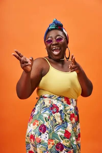 Возбужденный африканский американец плюс размер женщины с платком и солнцезащитные очки позируя на оранжевом фоне — стоковое фото