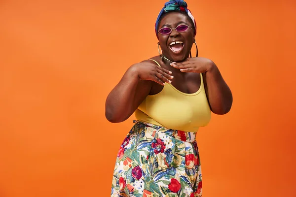 Excited african american plus size woman with headscarf and sunglasses posing on orange background — Stock Photo