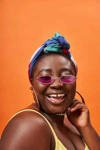Cheerful and plus size african american woman in headscarf and trendy sunglasses on orange — Stock Photo