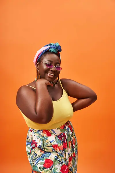 Positive african american plus size woman with headscarf and sunglasses posing on orange background — Stock Photo