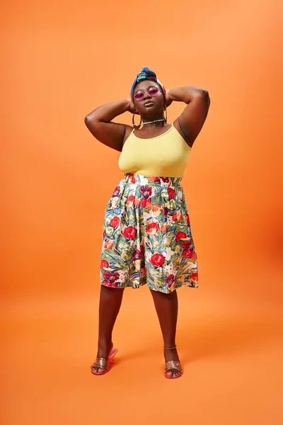 Stylish plus size african american woman in floral outfit and trendy sunglasses on orange backdrop — Stock Photo
