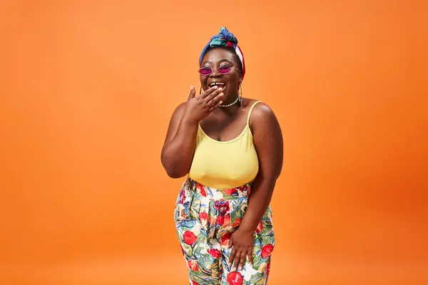 Happy plus size african american woman in floral outfit and sunglasses smiling on orange backdrop — Stock Photo