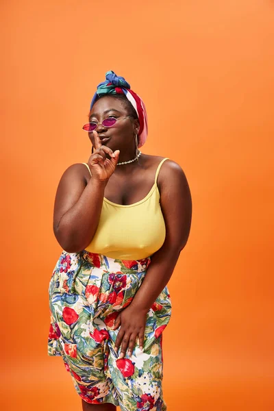 Plus size african american woman in floral outfit and sunglasses showing hush sign on orange — Stock Photo