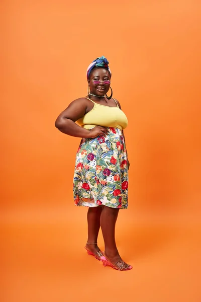 Plus size modelo americano africano em roupa floral e óculos de sol posando com a mão no quadril em laranja — Fotografia de Stock
