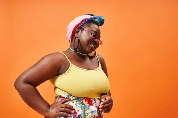Plus size african american woman in floral outfit and sunglasses posing with hand on hip on orange — Stock Photo