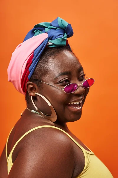 Joyeux plus la taille femme afro-américaine en foulard et lunettes de soleil à la mode sur fond orange — Photo de stock