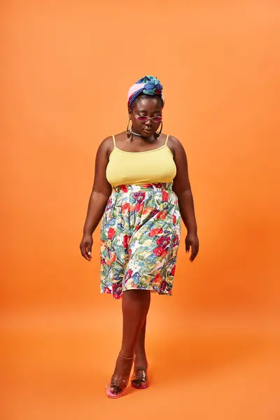 Plus size african american woman in floral outfit and stylish sunglasses walking on orange backdrop — Stock Photo