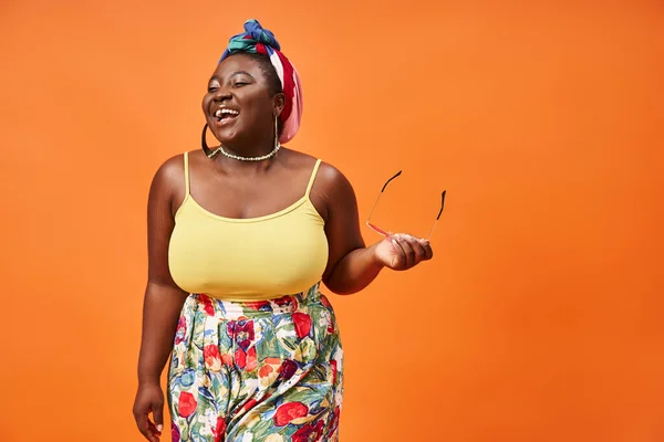 Happy plus size african american woman in floral outfit holding sunglasses on orange background — Stock Photo