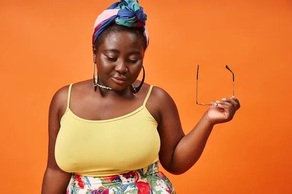 Plus size african american woman in floral outfit holding trendy sunglasses on orange background — Stock Photo