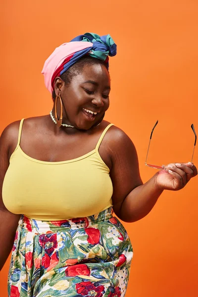 Smiling plus size african american woman in floral outfit holding sunglasses on orange backdrop — Stock Photo