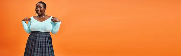 Heureuse femme afro-américaine taille plus en jupe à carreaux et bleu manches longues posant sur orange, bannière — Photo de stock