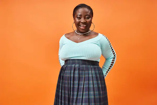 Body positive african american woman in plaid skirt and blue long sleeve smiling on orange backdrop — Stock Photo