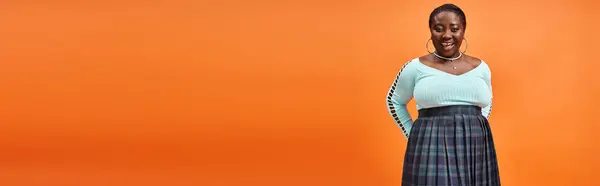 Corps positif femme afro-américaine en jupe à carreaux et bleu manches longues souriant sur orange, bannière — Photo de stock
