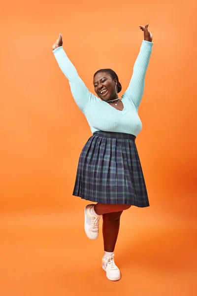 Alegre modelo afroamericano de talla grande en falda a cuadros y jersey de color menta posando en naranja - foto de stock