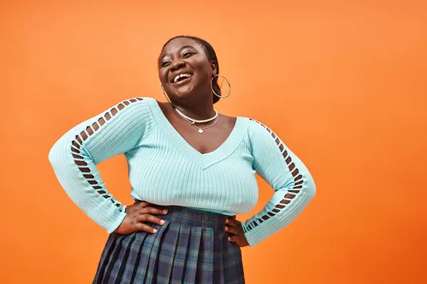Cheerful plus size african american woman in blue long sleeve posing with hands on hips on orange — Stock Photo