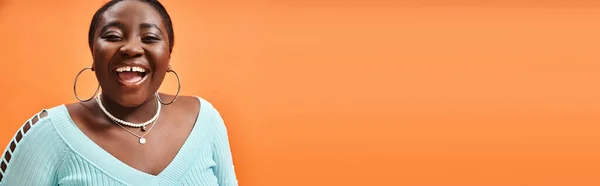 Portrait of cheerful plus size african american woman in blue long sleeve laughing on orange, banner — Stock Photo