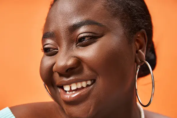 Portrait of cheerful plus size african american woman in blue long sleeve smiling on orange — Stock Photo