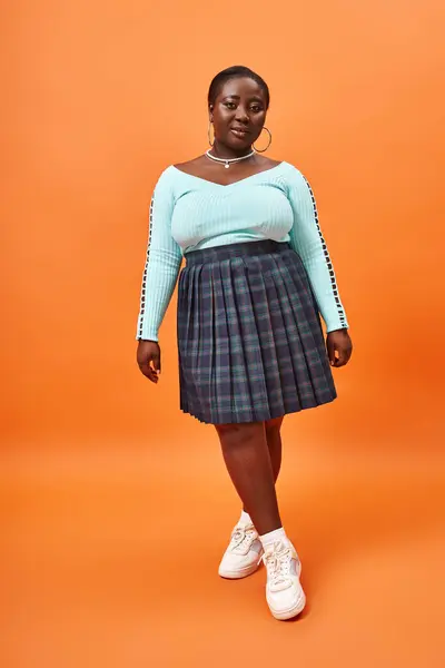 Comprimento total, além de tamanho mulher americana africana em saia xadrez e manga comprida posando em laranja — Fotografia de Stock
