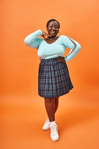 Feliz plus size mulher americana africana em saia xadrez e manga comprida sorrindo no fundo laranja — Fotografia de Stock