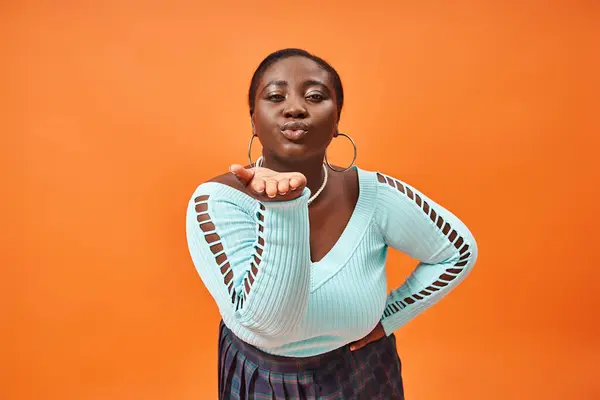 Plus size african american woman in plaid skirt and long sleeve sending an air kiss on orange — Stock Photo