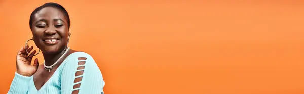 Portrait de femme africaine américaine gaie et de taille plus en bleu manches longues souriant sur orange, bannière — Photo de stock