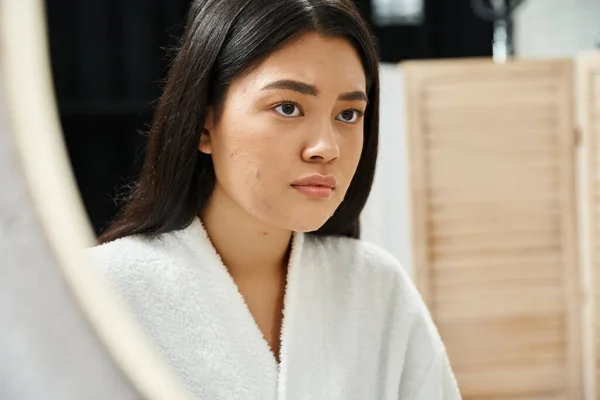Traurige asiatische Frau mit brünetten Haaren untersucht ihr Gesicht mit Akne im Badezimmerspiegel, Hautproblem — Stockfoto