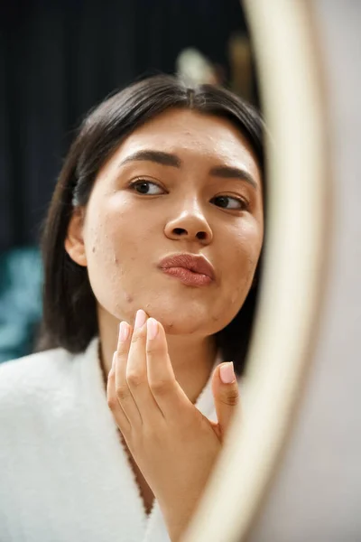 Jeune fille asiatique avec des cheveux bruns examiner son visage avec de l'acné dans le miroir de salle de bains, problème de peau — Photo de stock