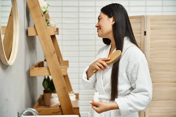 Young asian woman in robe combing her brunette hair and looking at bathroom mirror, side view — Stock Photo