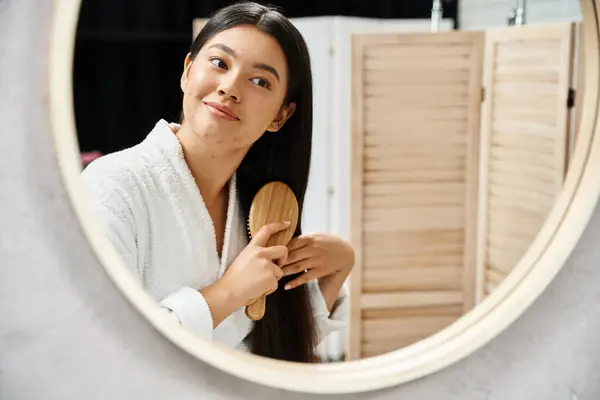 Joven asiático mujer en bata peinando su morena sano cabello y mirando baño espejo - foto de stock