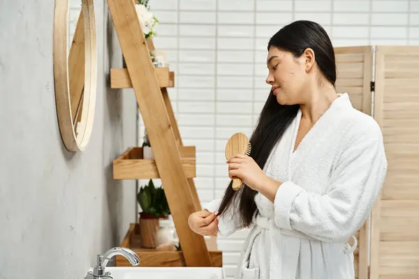 Side view of brunette and asian young woman in robe combing her brunette hair near bathroom mirror — Stock Photo
