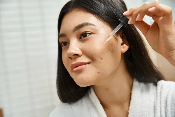 Joven y morena mujer asiática en bata de baño aplicando suero facial para tratar manchas en la cara - foto de stock