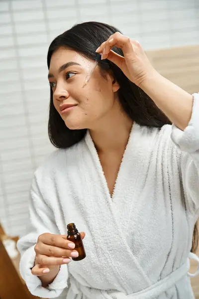 Smiling and young asian woman in bath robe applying facial serum to treat acne on face near mirror — Stock Photo