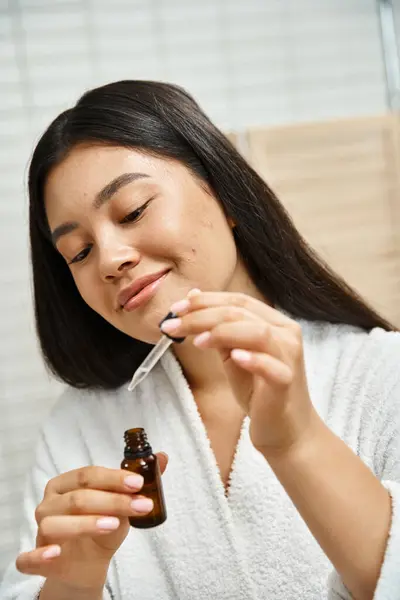 Joyful and asian woman in bath robe holding dropper with facial serum to treat acne on face — Stock Photo