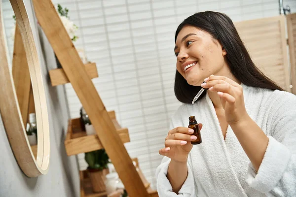 Fröhliche und junge asiatische Frau im Bademantel Anwendung Gesichtsserum zur Behandlung von Akne im Gesicht in der Nähe des Spiegels — Stockfoto