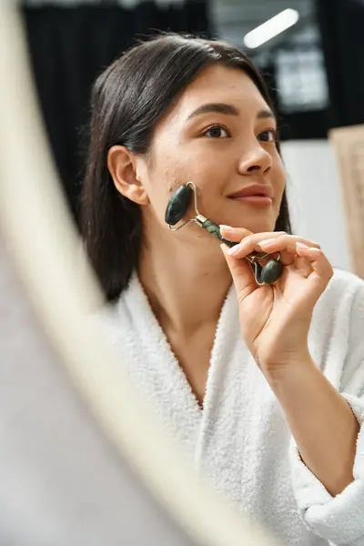 Smiling asian woman with brunette hair using jade roller for a facial massage in bathroom, skin care — Stock Photo