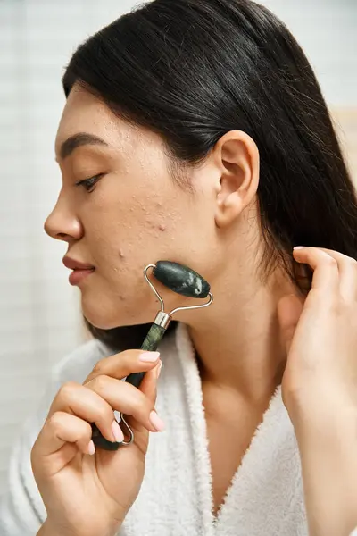 Close up photo of young asian woman with acne doing face massage with jade roller in bathroom — Stock Photo