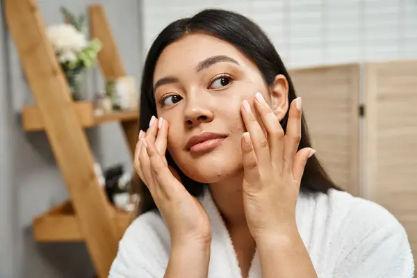 Conceito de cuidados com a pele, jovem mulher asiática em roupão de banho aplicando hidratante no rosto e olhando para longe — Fotografia de Stock