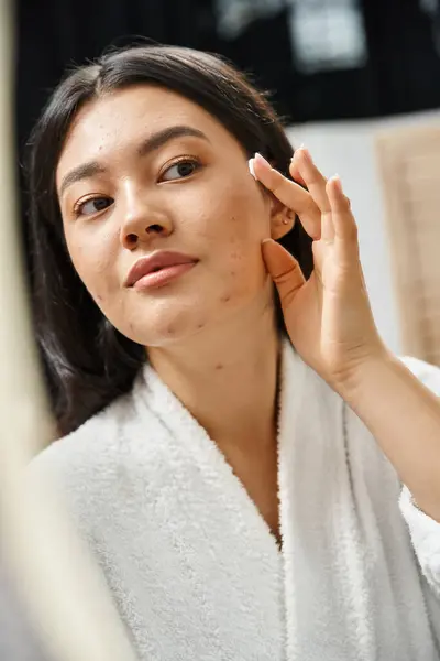 Primer plano de la mujer asiática con acné aplicación de crema en la cara y mirando el espejo en el baño - foto de stock