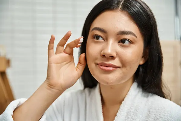 Gros plan de jeune femme asiatique avec l'acné appliquer la crème sur le visage et regarder miroir dans la salle de bain — Photo de stock
