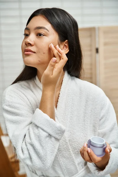 Brunette asian woman with acne applying cream on face and looking away in bathroom, skin issues — Stock Photo