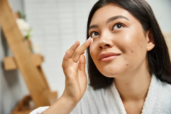 Morena asiática mulher com acne aplicação de creme no rosto e olhando para cima no banheiro, problemas de pele — Fotografia de Stock