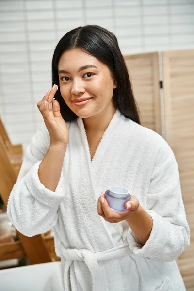 Happy asian woman with acne applying cream on face and smiling while looking at camera in bathroom — Stock Photo