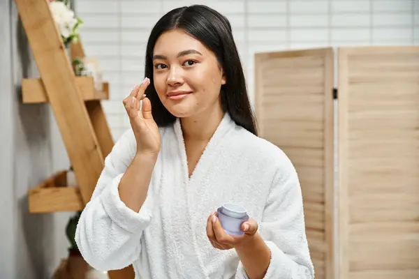 Cheerful asian woman with acne applying cream on face and smiling while looking at camera at home — Stock Photo