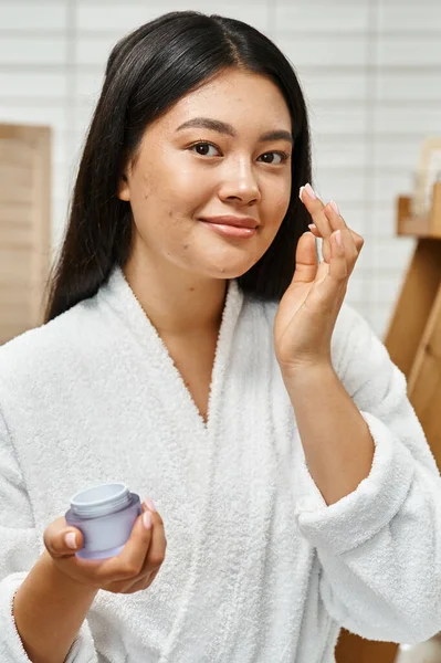 Cheerful asian woman with acne applying cream on face and smiling while looking at camera, vertical — Stock Photo