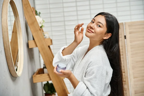Cheerful and pretty asian woman with acne applying cream on face and smiling while looking at camera — Stock Photo