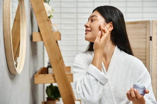 Gai et jolie asiatique femme avec acné application crème sur le visage et en regardant salle de bain miroir — Photo de stock